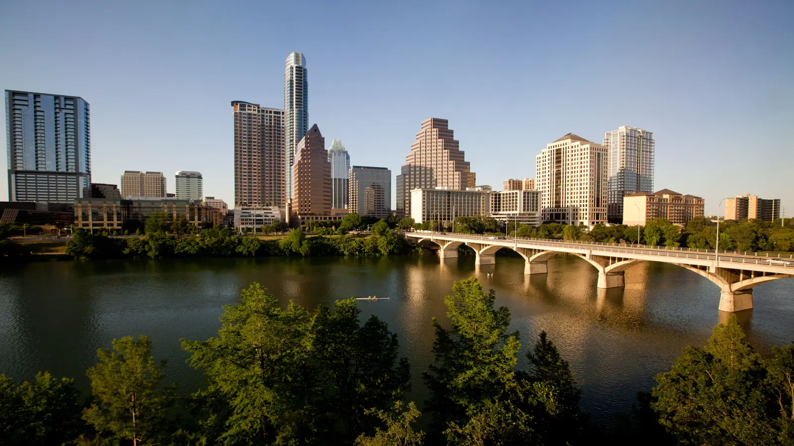 Downtown Austin at sunset above Lady Bird Lake and the Congress Avenue Bridge, framed by trees.