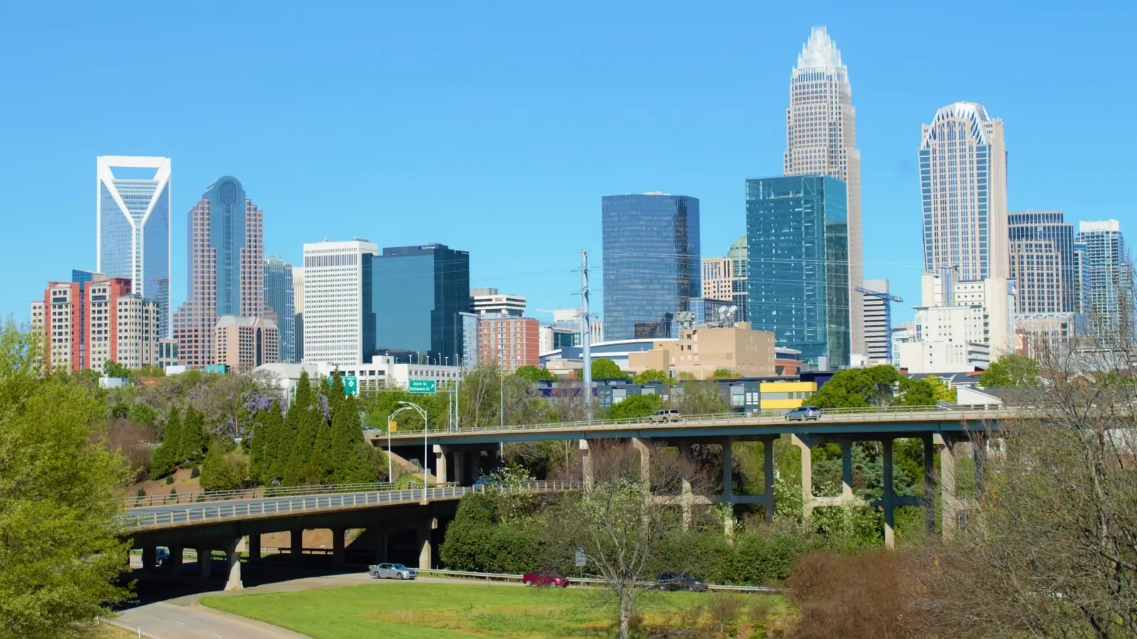 Uptown Charlotte skyscrapers behind a park footbridge under a blue sky.