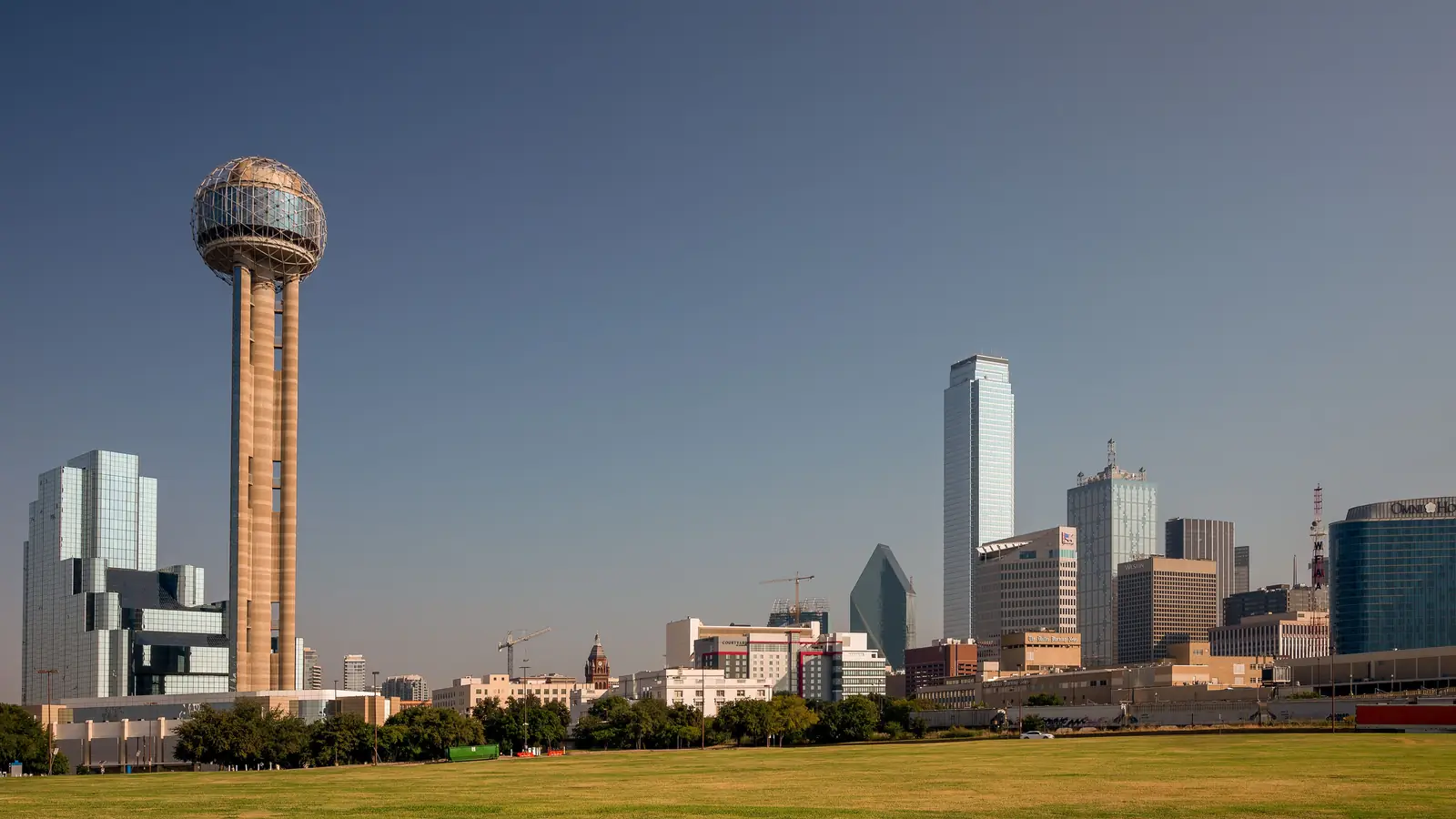 Reunion Tower and the downtown Dallas skyline under a calm sky, with an open lawn in the foreground.