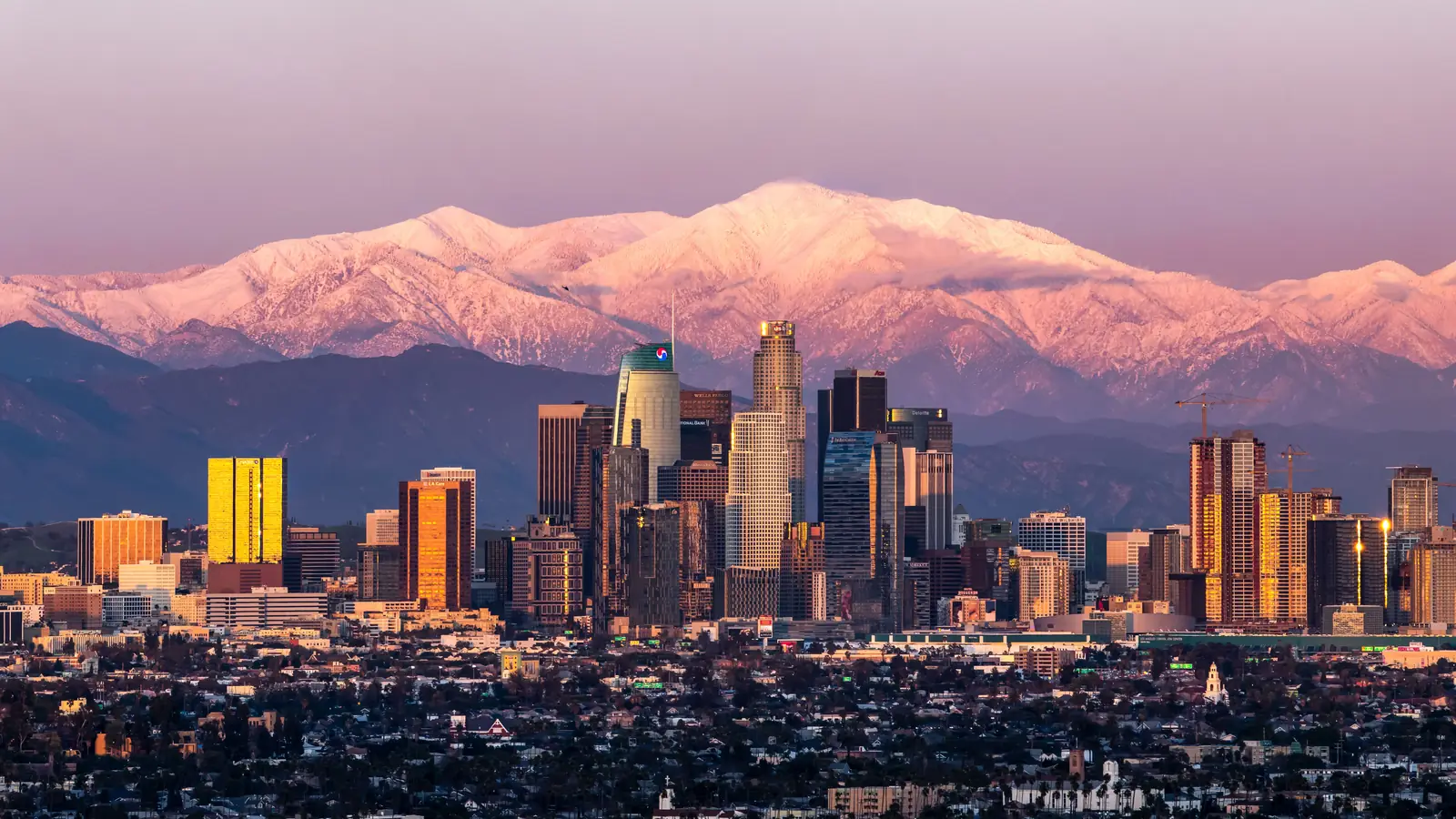Downtown Los Angeles at sunset, with snow-covered Mount Baldy and the San Gabriel Mountains glowing pink behind the skyline.