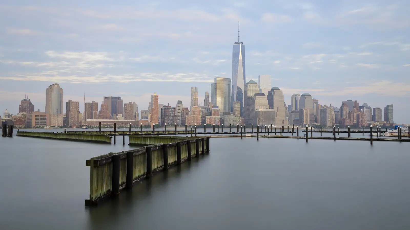 Lower Manhattan and One World Trade Center in New York across the calm Hudson River, seen from a pier in Jersey City.